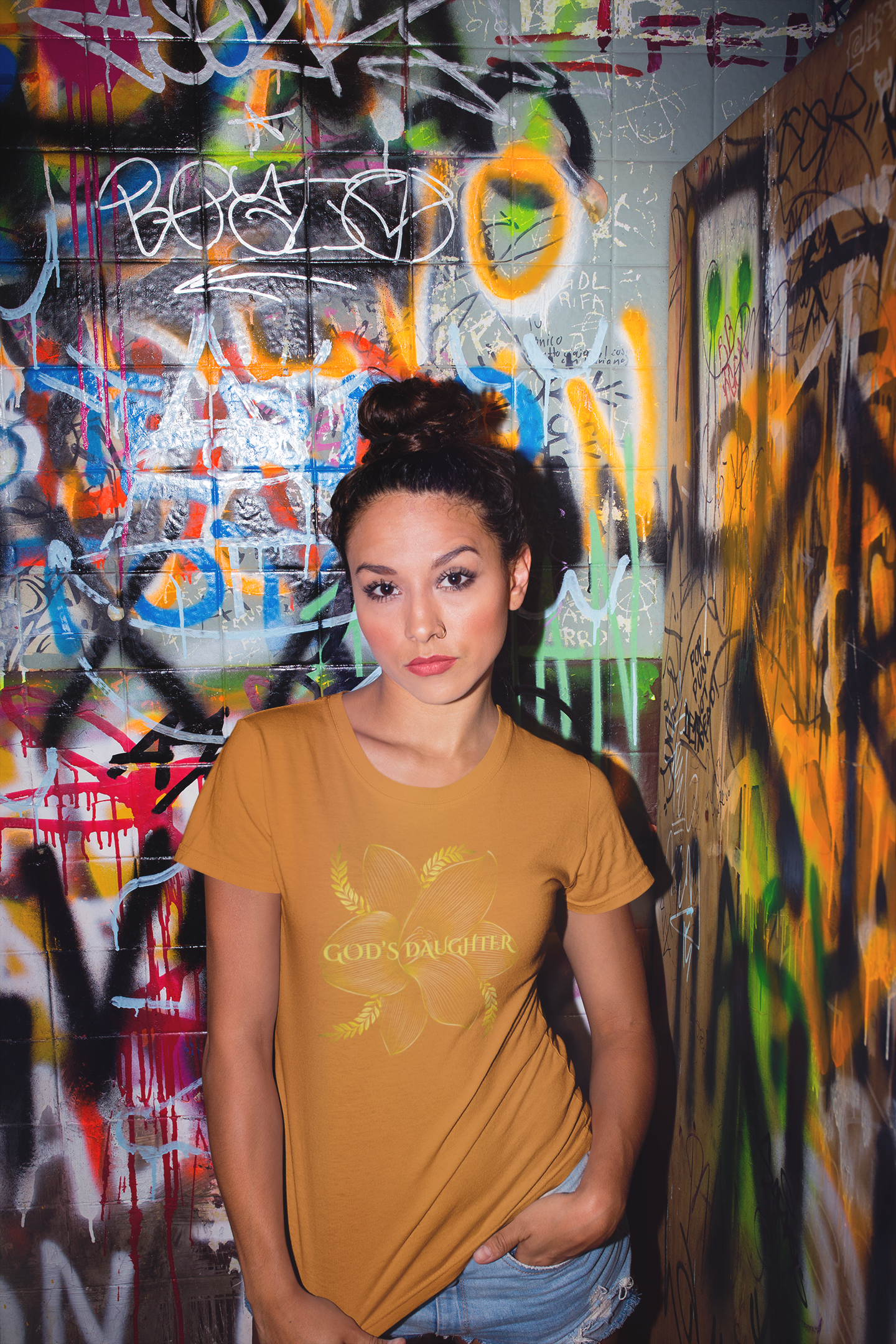 Woman wearing a mustard yellow t-shirt with text in front of a graffiti-covered wall.